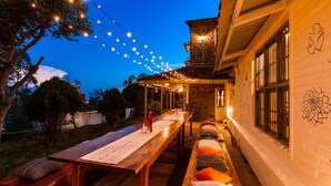 Un espacio de comedor al aire libre con una larga mesa de madera, bancos y luces colgantes.