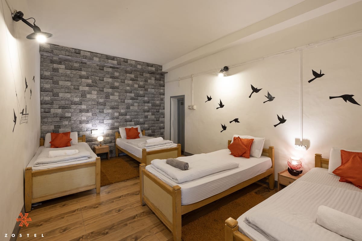 A room with three wooden beds, each with white linens and orange pillows. A stone wall and a mural of birds are on the walls.