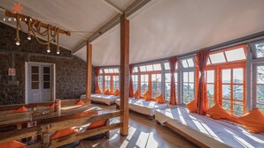 Una habitación rústica con bancos y camas de madera, ventanas grandes con cortinas naranjas y una pared de piedra.