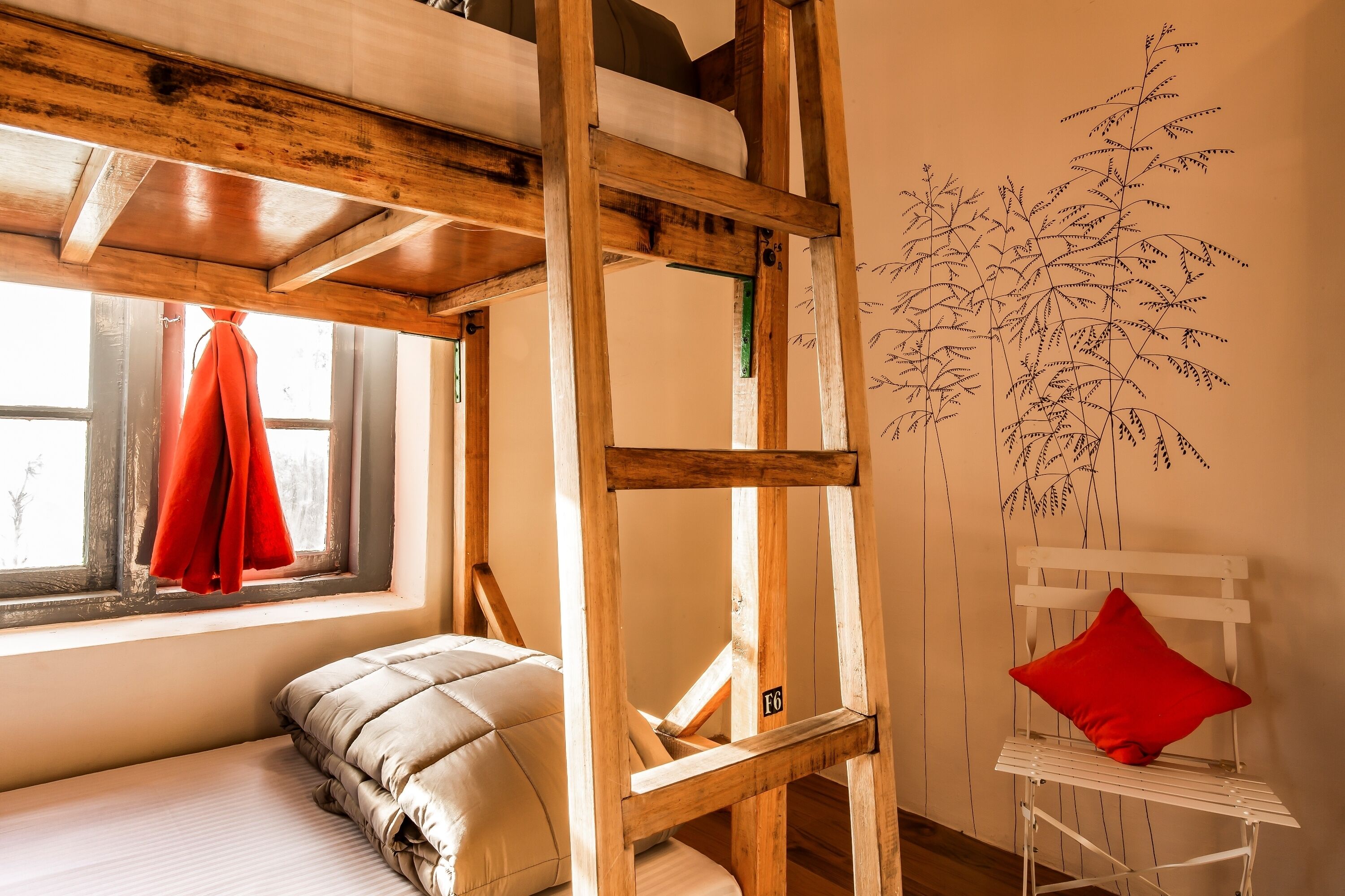 Una habitación con cama de altillo, una escalera de madera, una ventana con cortina roja, una silla blanca con cojín rojo y un mural en la pared.