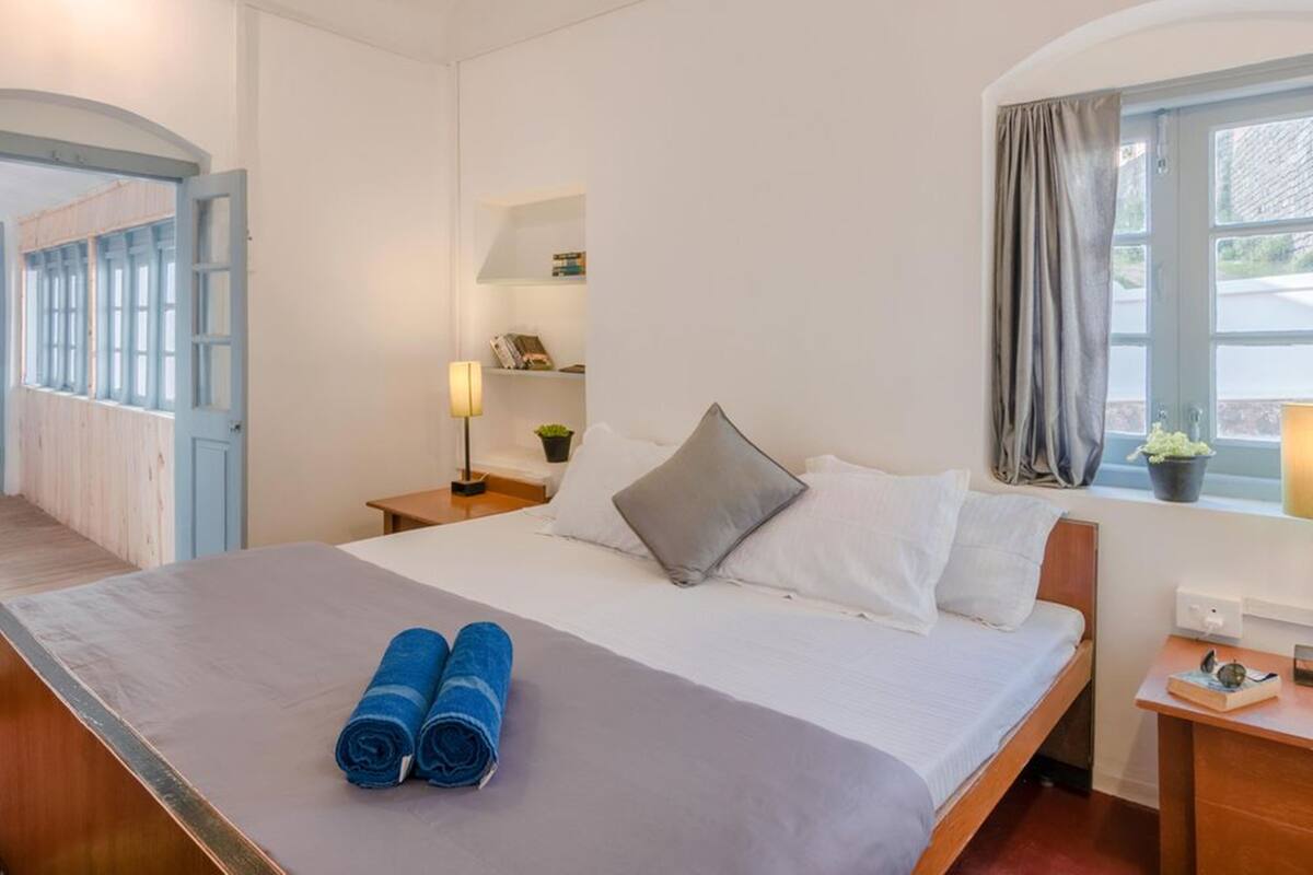 A bedroom with a bed, pillows, a nightstand, a lamp, a window with curtains, and a view of the outside.