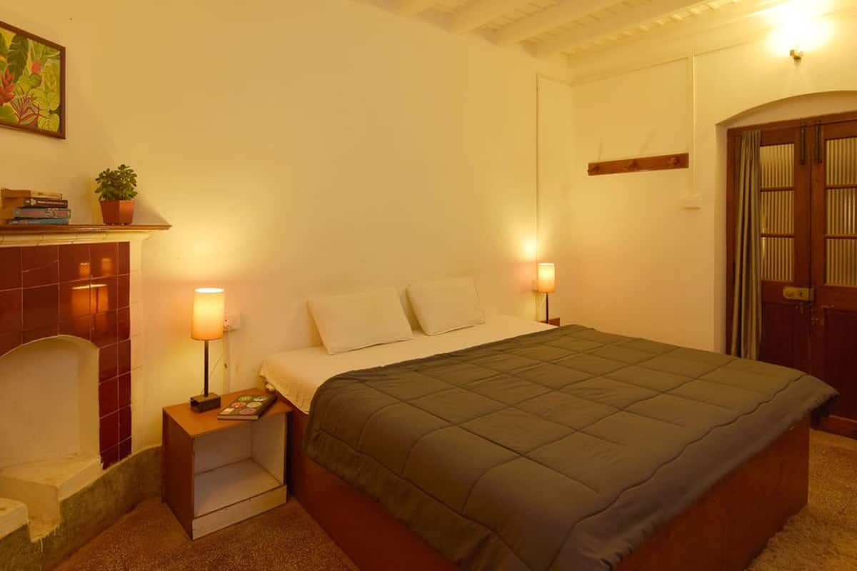A bedroom with a bed, bedside lamps, a nightstand, a small table with books, a potted plant, and a painting on the wall.
