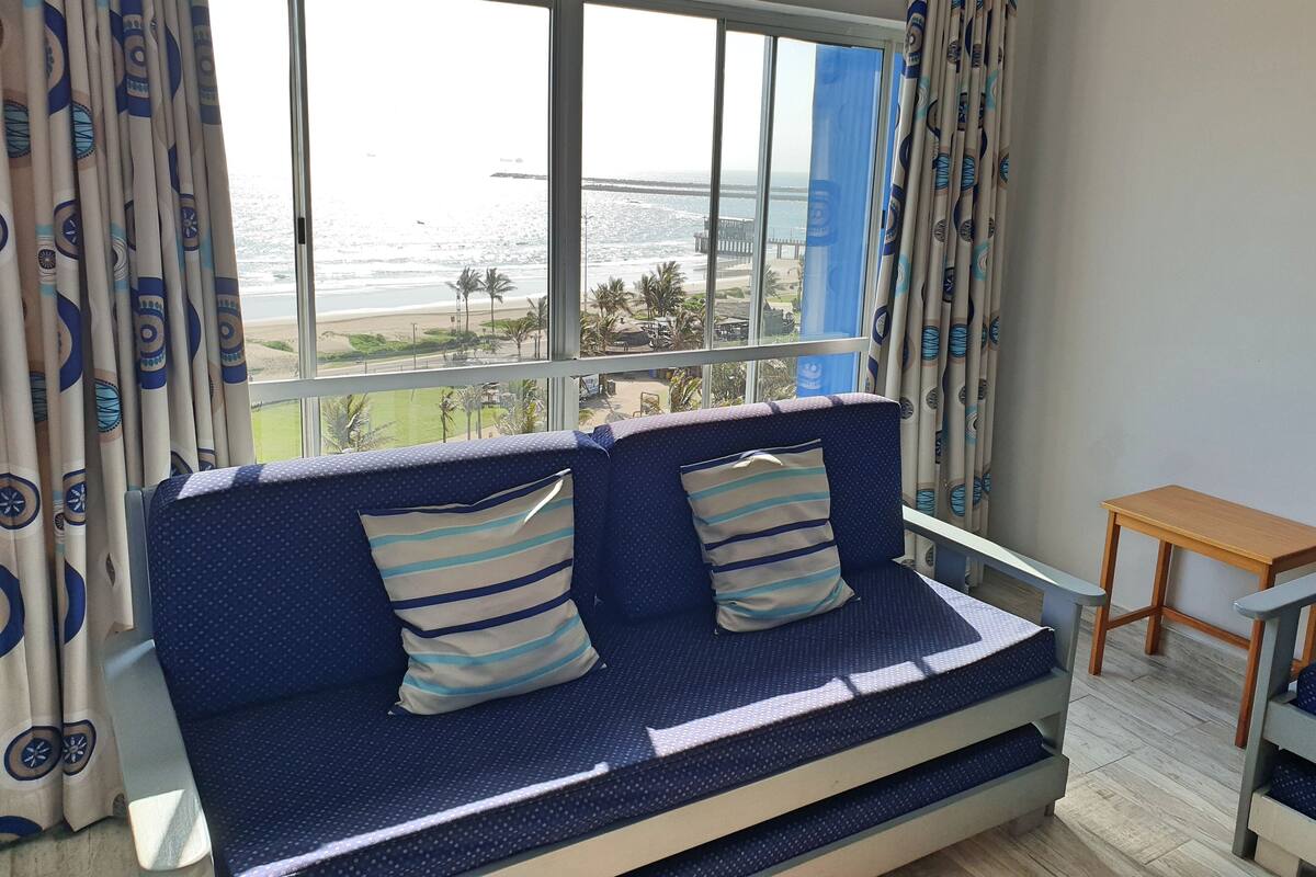 A room with a large window offering a view of the beach and ocean. There is a blue sofa with striped pillows and a small wooden table.