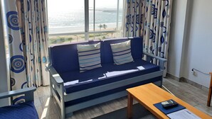 A room with a sofa facing a large window, a wooden table, and a view of the sea. - Silver Sands Self Catering (Durban)