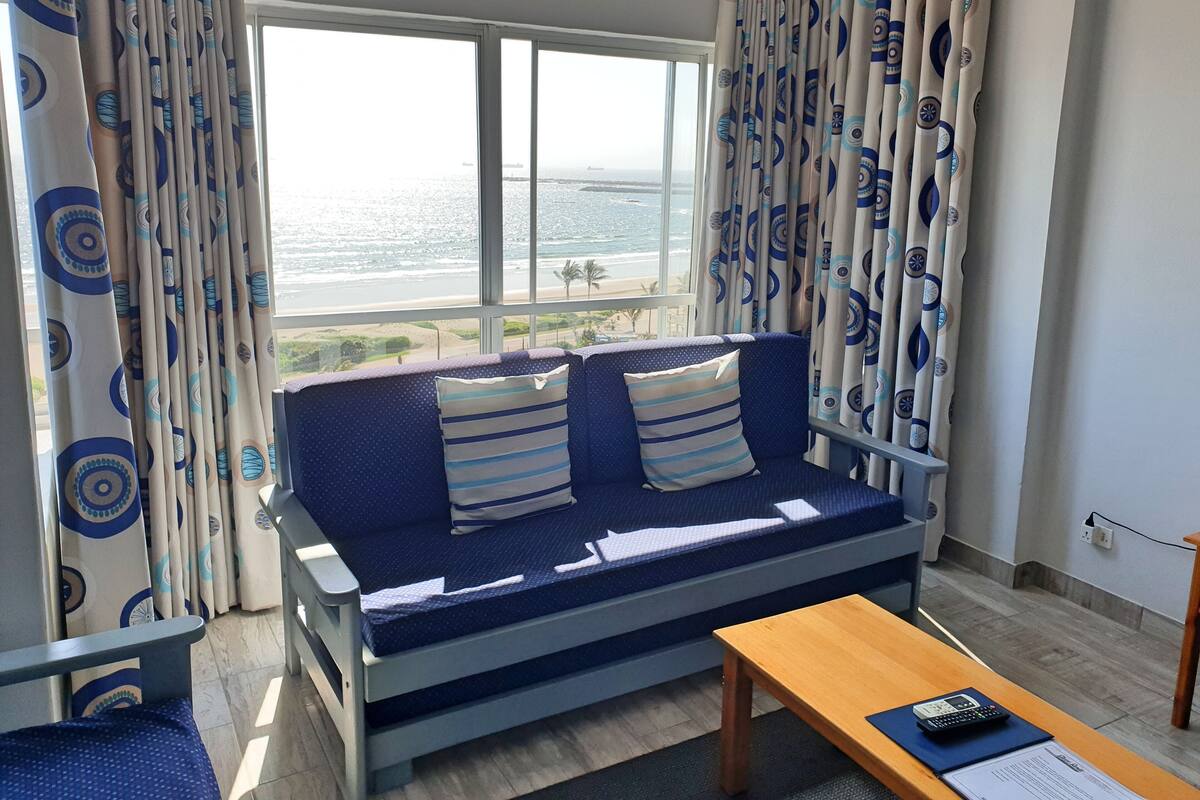 A room with a sofa facing a large window, a wooden table, and a view of the sea.
