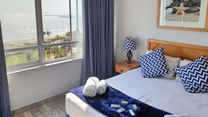 A hotel room with a large window offering a view of the beach and ocean. There is a bed with blue and white pillows, a nightstand with a lamp, and a framed picture on the wall. - Silver Sands Self Catering (Durban)