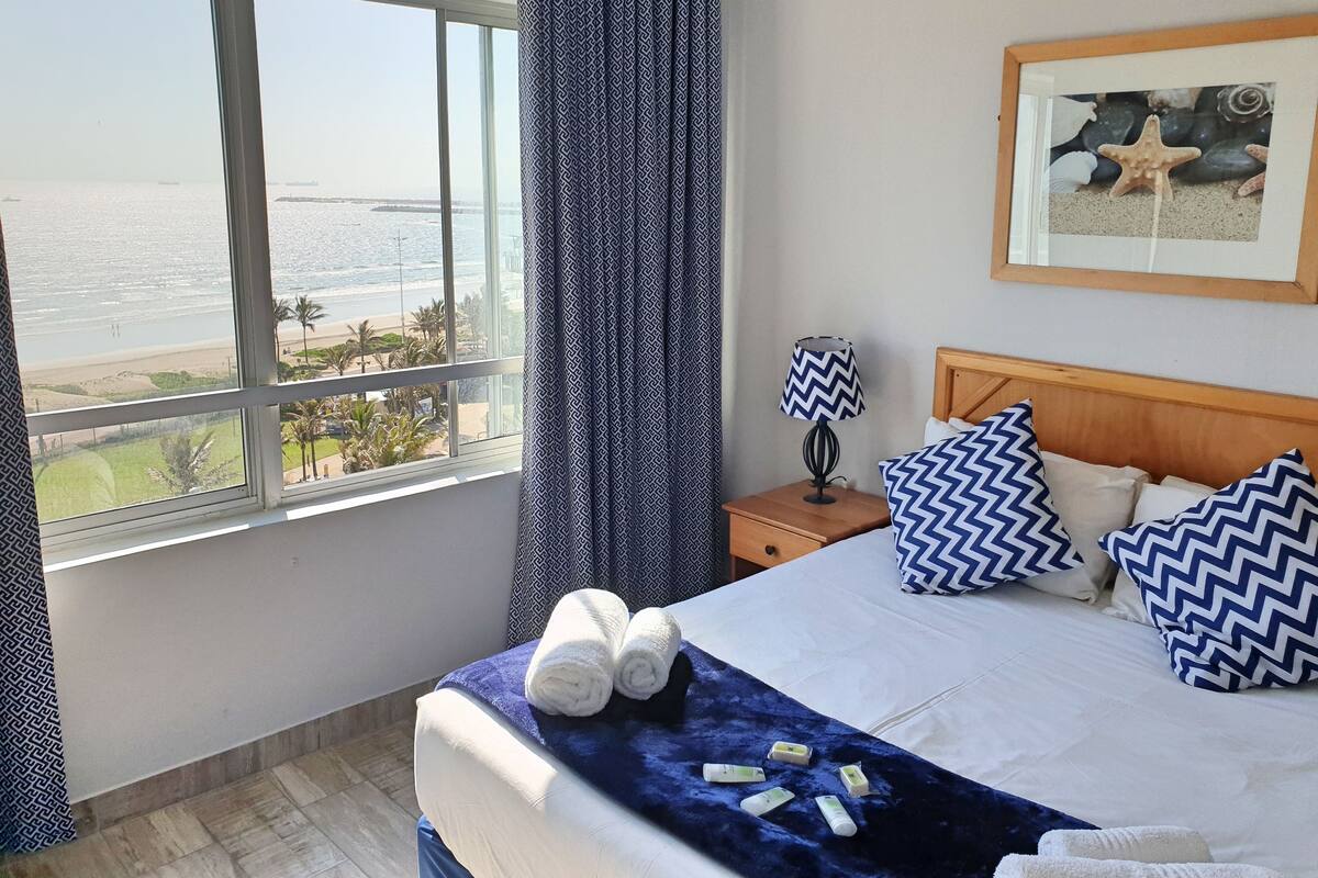 A hotel room with a large window offering a view of the beach and ocean. There is a bed with blue and white pillows, a nightstand with a lamp, and a framed picture on the wall.