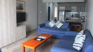 A living room with a sofa, a coffee table, and a TV. There is a kitchen area visible in the background. - Silver Sands Self Catering (Durban)