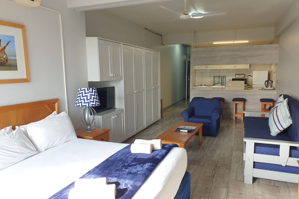A hotel room with a bed, a desk, a chair, a sofa, a TV, and a kitchenette.