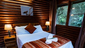 Un dormitorio de cabaña de madera con una cama, almohadas, una manta, una mesita de noche con una lámpara y una ventana con vistas a árboles.