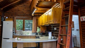 Interior rústico de una cabaña con techo de madera, una zona de cocina que incluye un refrigerador, estufa y armarios de madera, y una barra con taburetes.