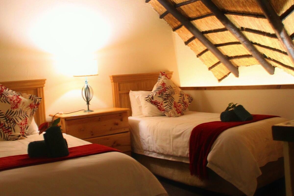 A room with two beds, each with floral-patterned pillows and a red blanket. There is a nightstand with a lamp between the beds.