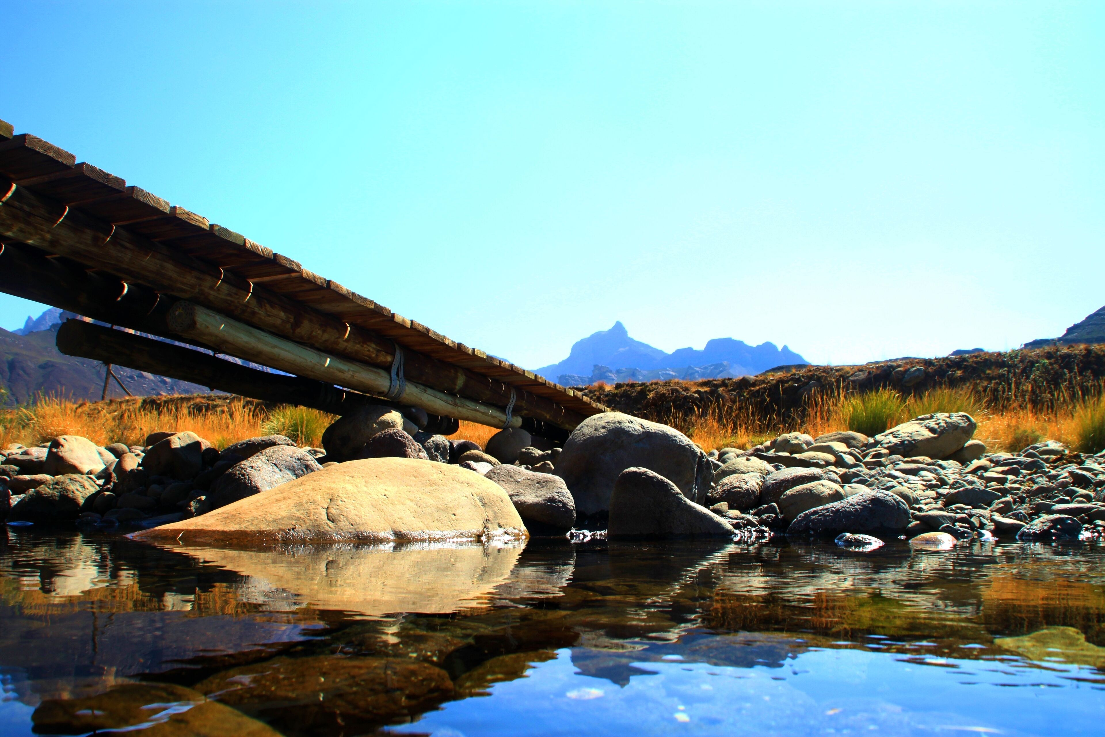 A wooden bridge over a calm river with large rocks, clear water, and mountains in the background.
