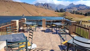 Une terrasse extérieure avec des tables et des chaises, offrant une vue sur un plan d’eau et des montagnes.