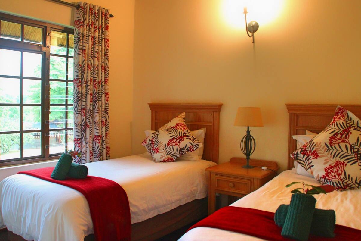 A hotel room with two beds, a window with curtains, a bedside table with a lamp, and a wall-mounted light fixture.