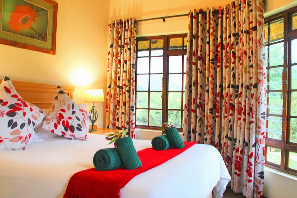 A hotel room with a bed, pillows, a red blanket, rolled towels, a window with curtains, and a painting on the wall.