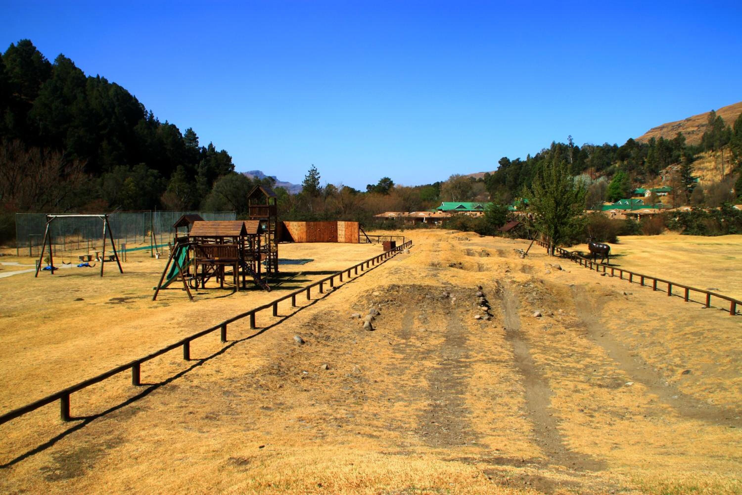 A fenced outdoor area with a wooden play structure, a dirt path, and a small building.