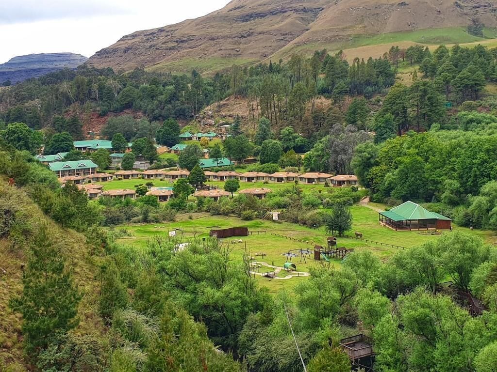 A rural setting with multiple green-roofed buildings, likely a guesthouse or lodge, surrounded by lush greenery and hills in the background.