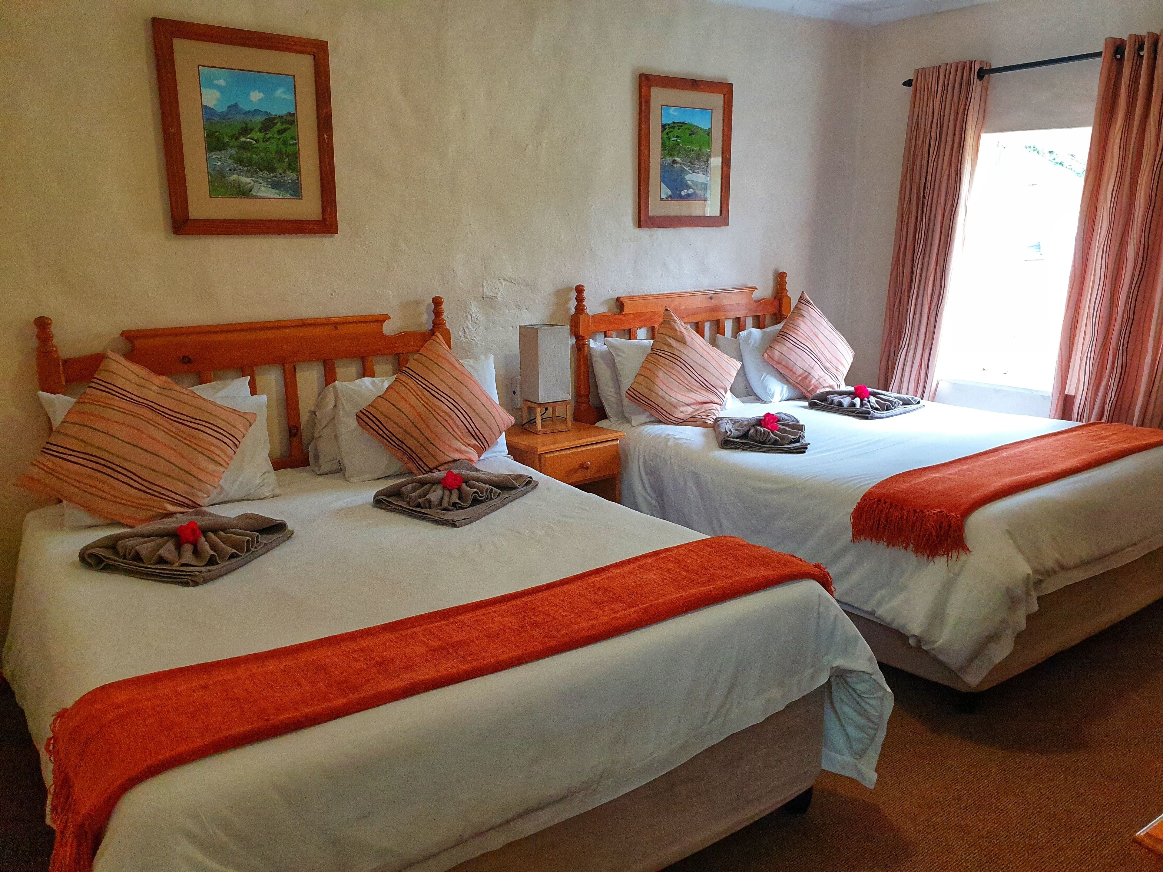 Ein Hotelzimmer mit zwei Betten, jeweils mit einem Holz-Kopfteil, weißen Bettwäsche und gestreiften Kissen. An den Wänden hängen zwei gerahmte Bilder, ein Fenster mit Vorhängen und ein kleiner Tisch mit einem Lautsprecher und einem gefalteten Handtuch.