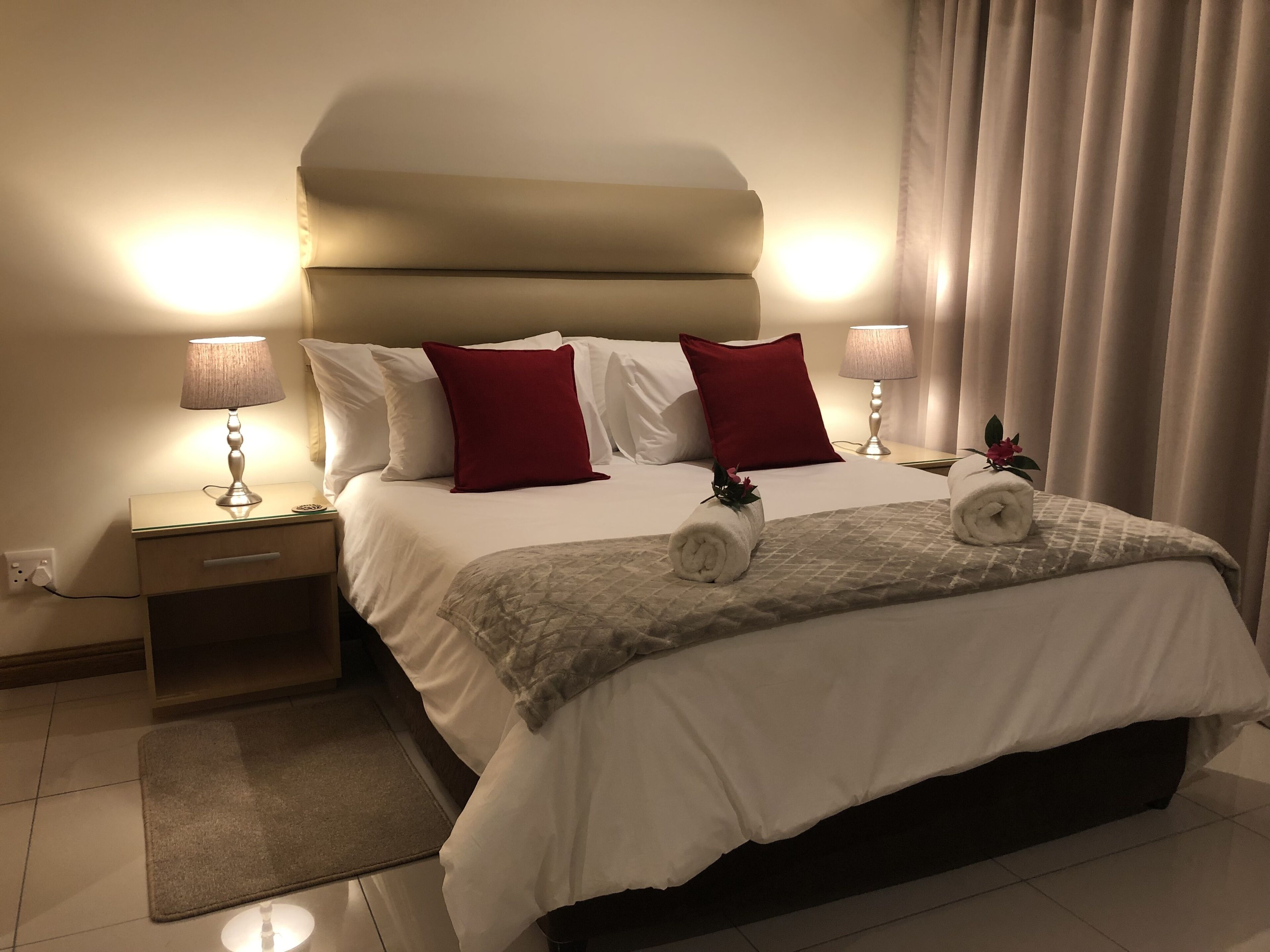Una cama bien hecha con almohadas blancas y rojas, una toalla enrollada y un edredón. Hay dos lámparas de noche, una mesita de noche y una pequeña mesa con una lámpara.