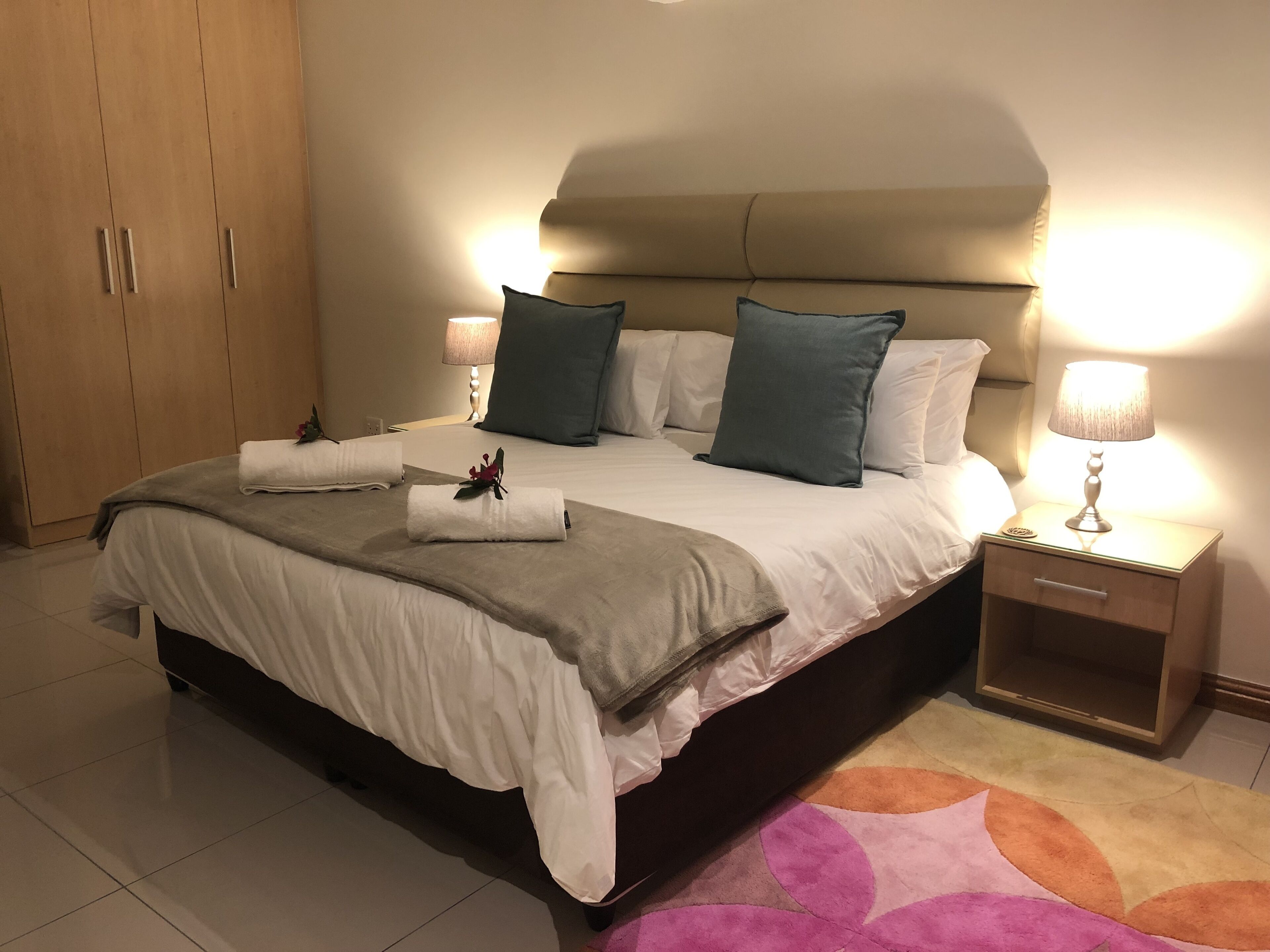 Una cama bien hecha con almohadas, una mesita de noche con lámpara y un armario de madera.