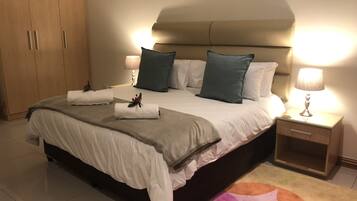 Una cama bien hecha con almohadas, una mesita de noche con lámpara y un armario de madera.
