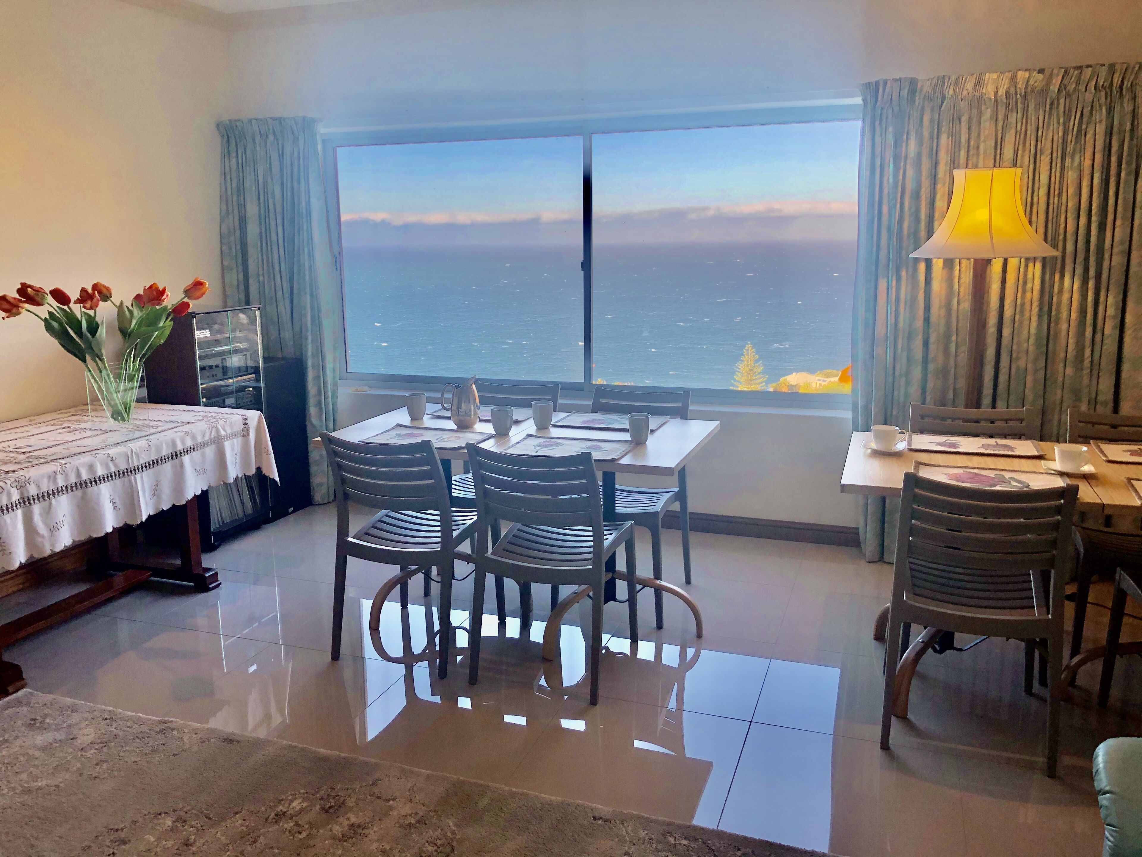 A dining area with a table set for four, a vase with flowers, and a view of the sea through large windows.