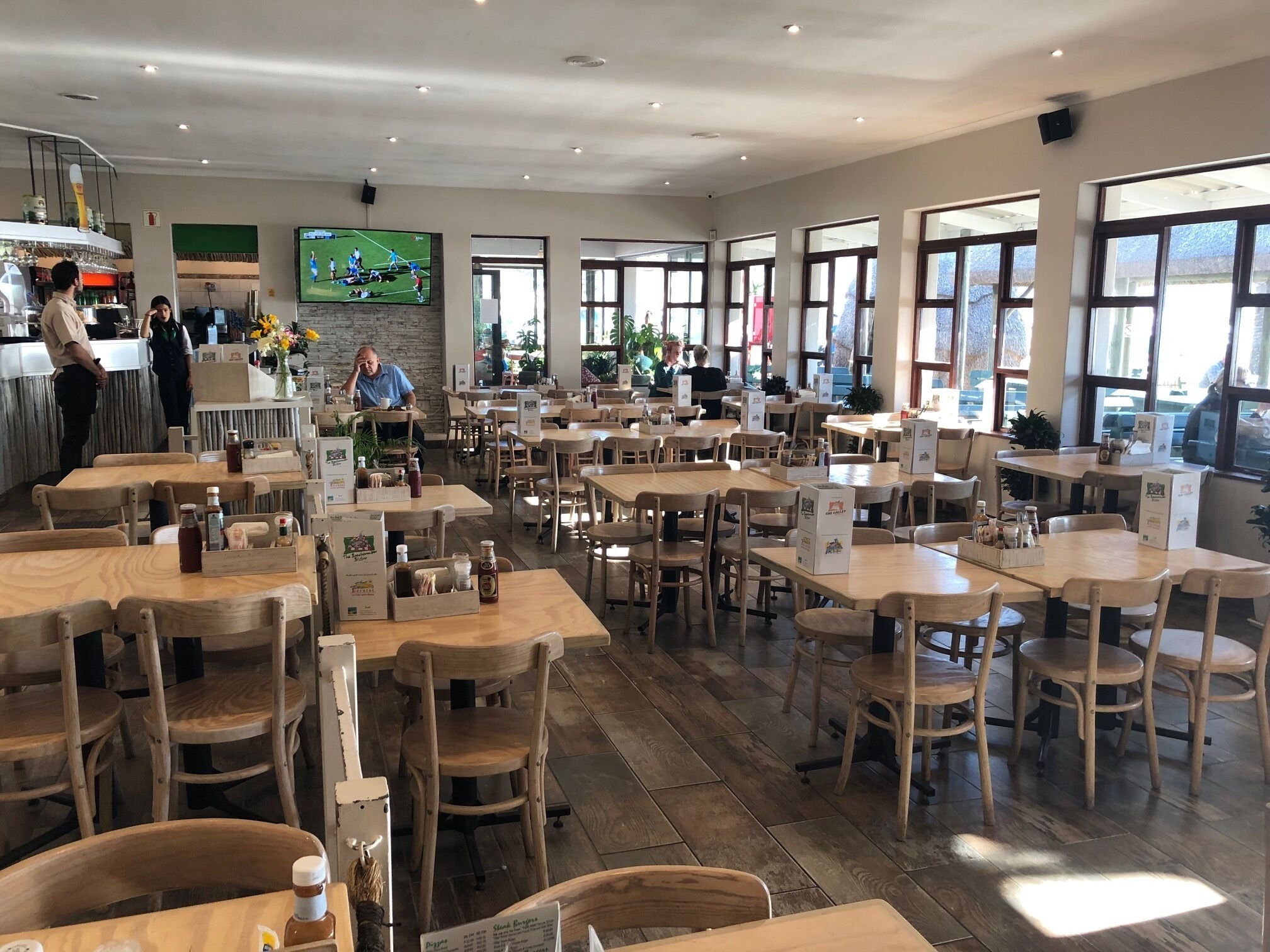 A spacious restaurant with wooden tables and chairs, a bar area, and large windows allowing natural light.