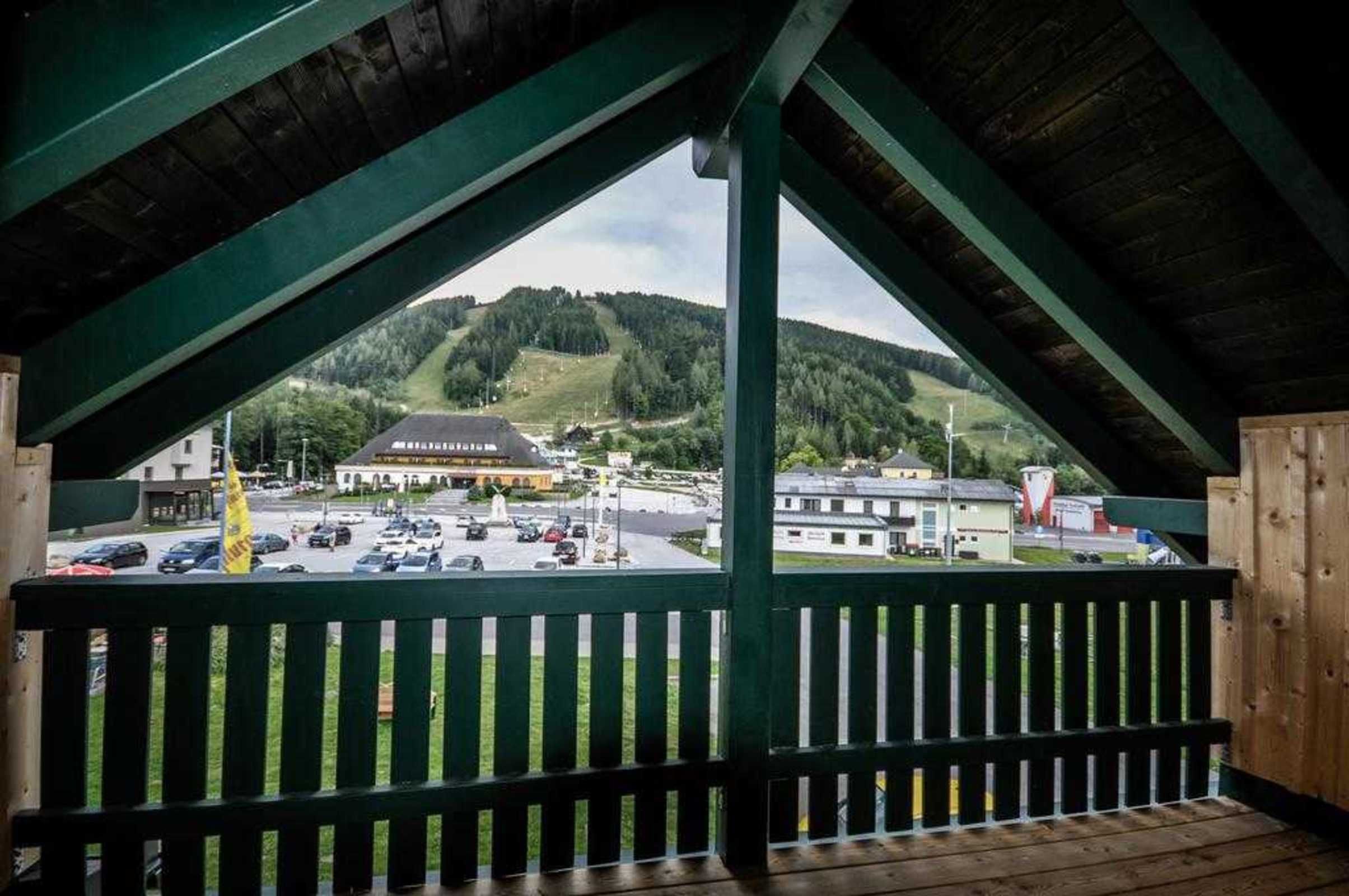 Uitzicht vanaf een overdekt balkon met houten leuning, op een parkeerplaats en gebouwen, waaronder een hotel met een kenmerkend dak.