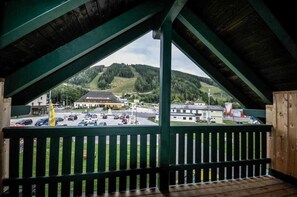 A view from a covered balcony with a wooden railing, overlooking a parking lot and buildings, including a hotel with a distinctive roof. - Styrian apartment with large balcony - Steirer Apartment (Semmering-Kurort)