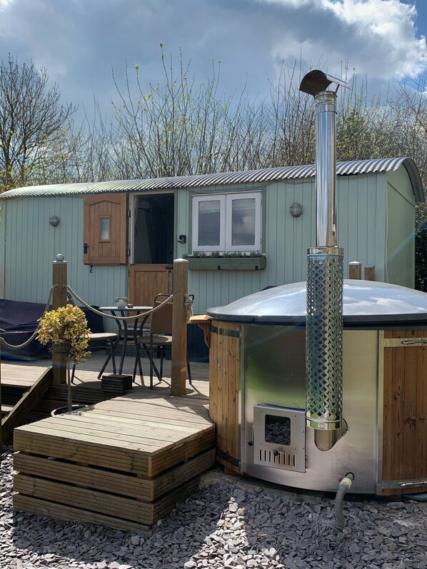 A rustic outdoor seating area with a wooden table and chairs, a small wooden structure with a window, and a metal grill station. - Large New Forest Shepherds Hut and Scandinavian Hot Tub with hydro massager <br><br> (Sway)