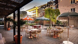 Un patio al aire libre con mesas y sillas, rodeado de edificios y luces colgantes.