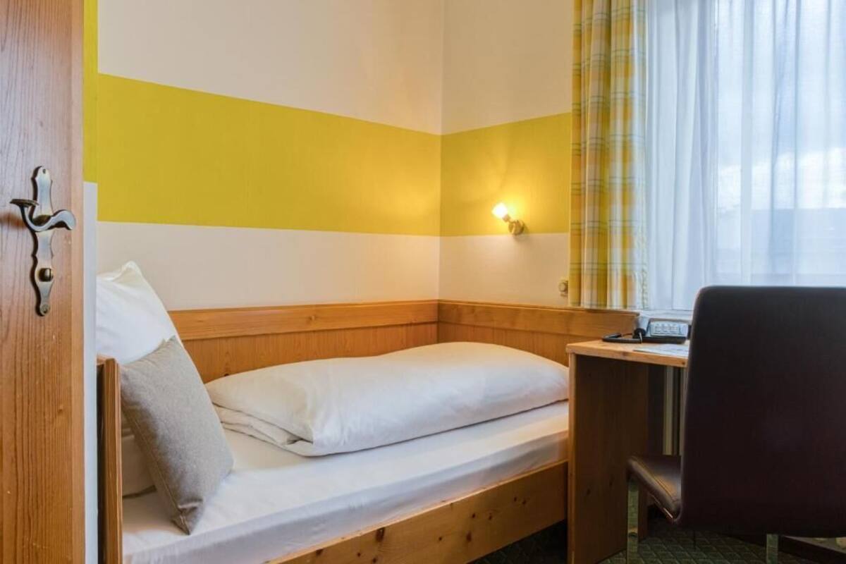 Ein kleines Zimmer mit einem Einzelbett, einem Holzkopfteil, einem Schreibtisch mit Stuhl und einem Fenster mit durchsichtigen Vorhängen.