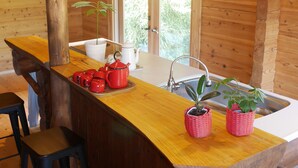A wooden cabin interior with a kitchen area featuring a wooden bar, stools, hanging pendant lights, and potted plants. - Shimano Log House (Iki)