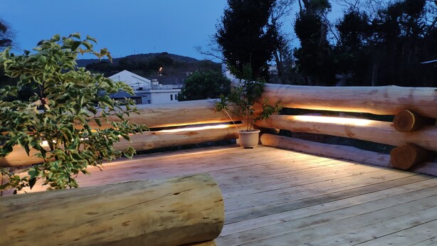 A wooden deck with a bench and potted plants, surrounded by trees and a building in the background. - Shimano Log House (Iki)