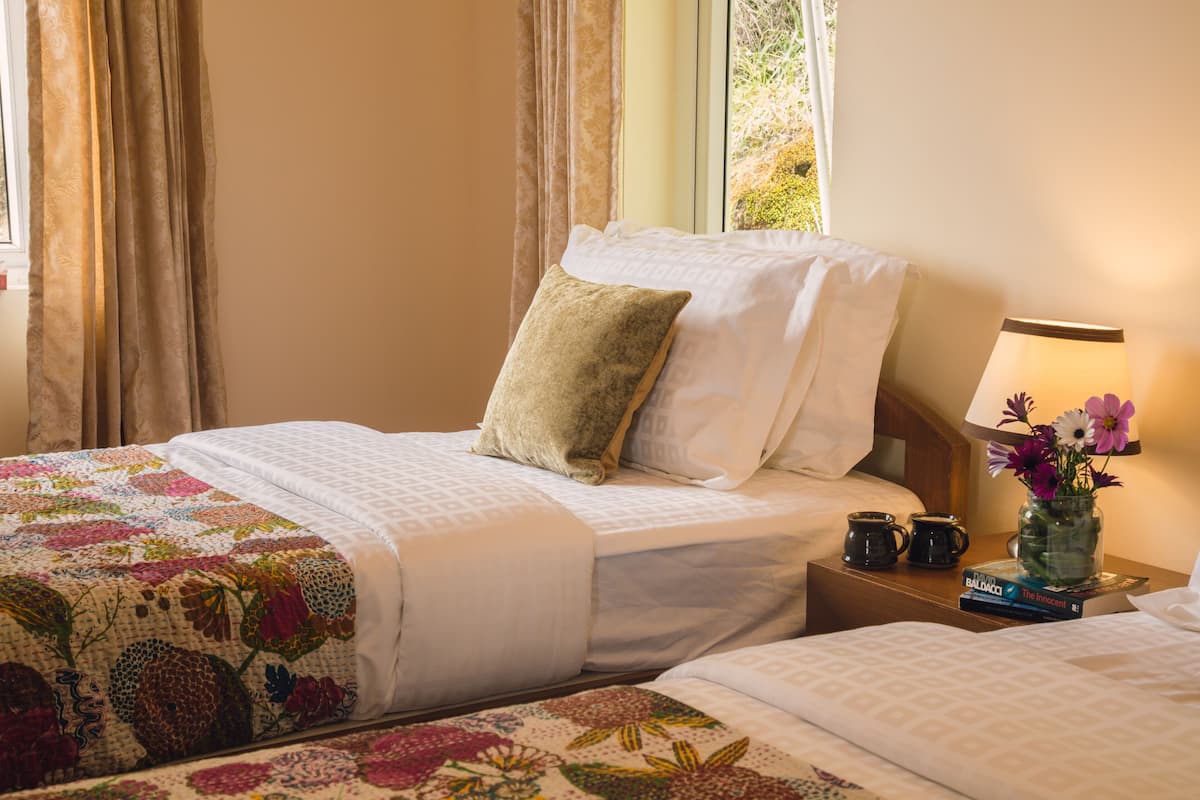 Habitación de hotel con dos camas, una mesita de noche con lámpara y un jarrón de flores, y una ventana con cortinas.