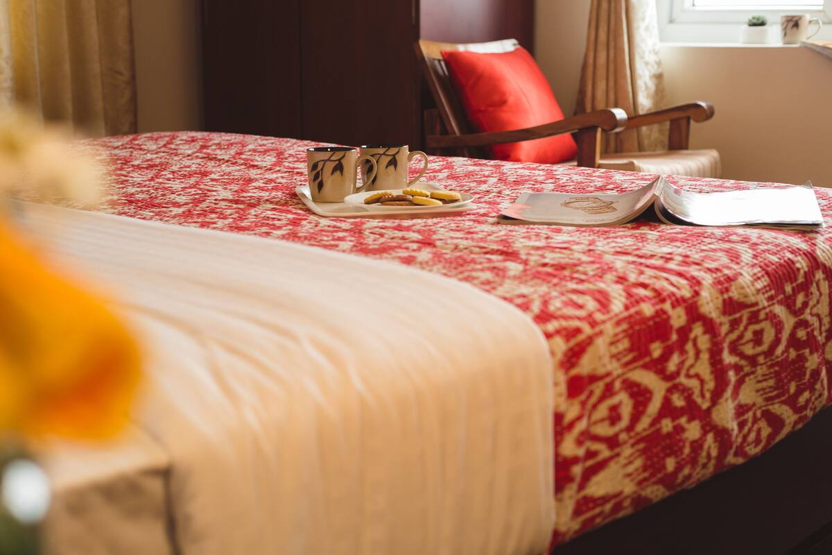Una cama bien hecha con colcha de cuadros rojos y blancos, dos tazas, un plato con galletas y un libro abierto sobre una bandeja.