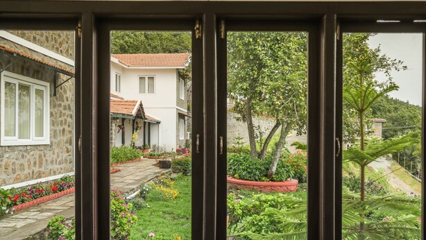 Vista a través de una ventana que muestra un jardín con un muro de piedra, un sendero y un edificio con techo de tejas rojas.