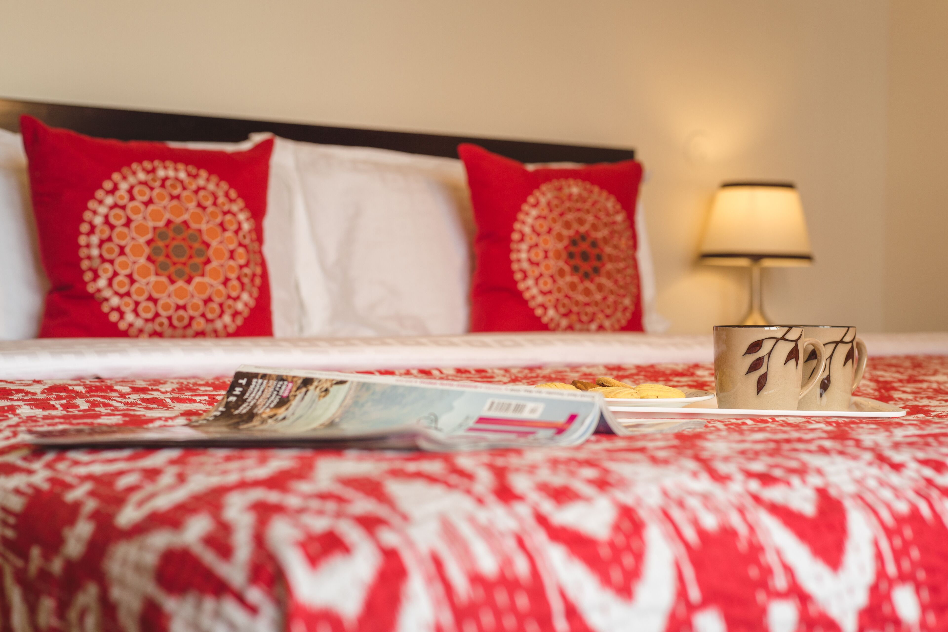 Una cama bien hecha con ropa de cama roja y blanca, una revista, una taza y una lámpara sobre una mesita de noche.