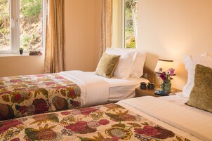 Una habitación con dos camas, una mesita de noche con lámpara, un jarrón con flores y una ventana con cortinas.