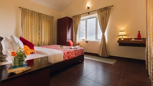 Habitación de hotel con cama, mesita de noche, armario, ventana con cortinas, una mesilla con lámpara y un estante pequeño con una botella.