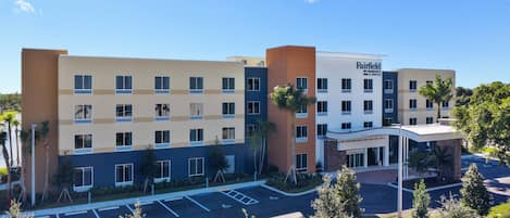 A multi-story hotel with a modern design, featuring a mix of beige and blue exterior colors, a central entrance, and a parking lot with marked spaces.
