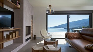 Een moderne woonkamer met een grote televisie, houten planken, een witte stoel, een leren bank en uitzicht op zee.