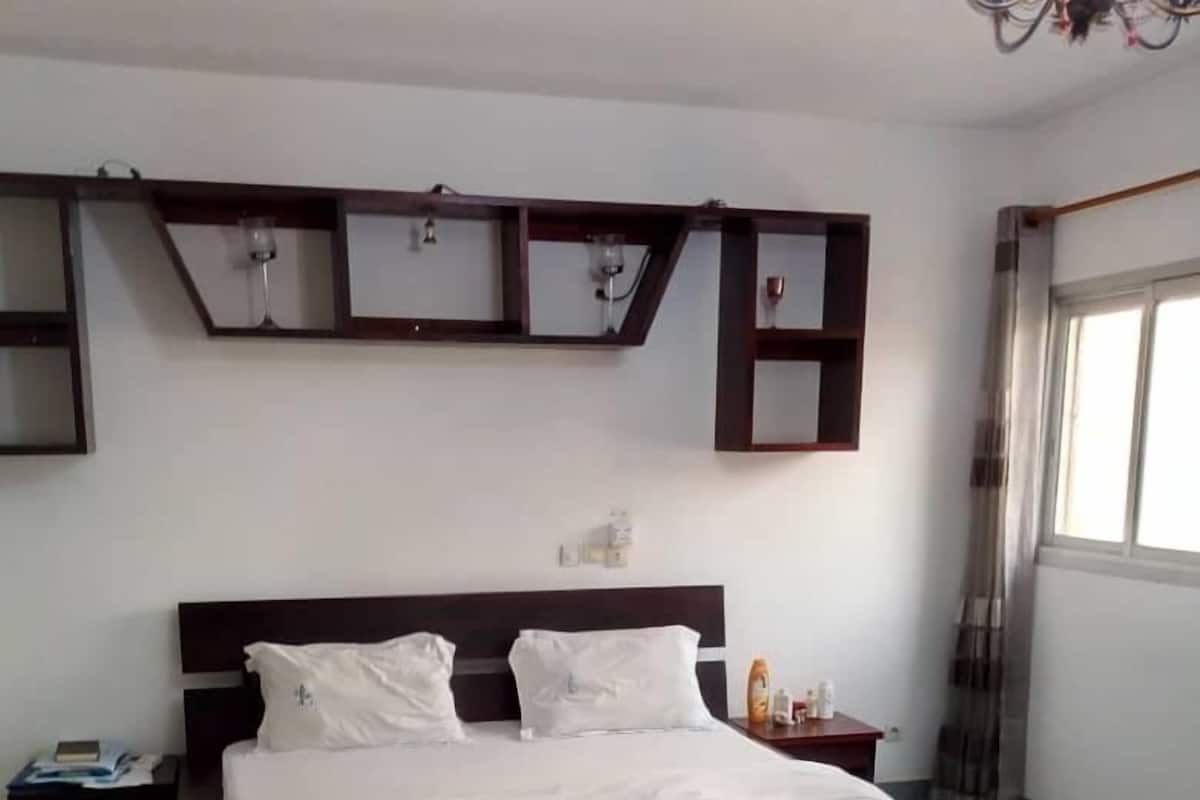 A neatly made bed in a hotel room with a large window, a chandelier, and a small bedside table with a bottle.