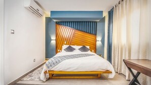 A modern bedroom with a wooden headboard, a bed with white bedding, a small table, and a curtain on the window. - Appartement Meublé VIP Douala Bonapriso (Douala)