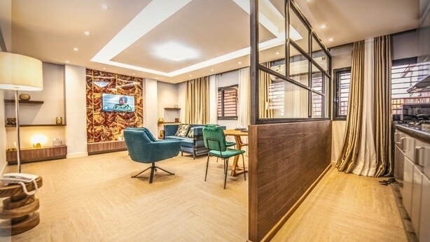 A modern hotel room with a dining area, a TV mounted on the wall, and a view through a glass partition. - Appartement Meublé VIP Douala Bonapriso (Douala)