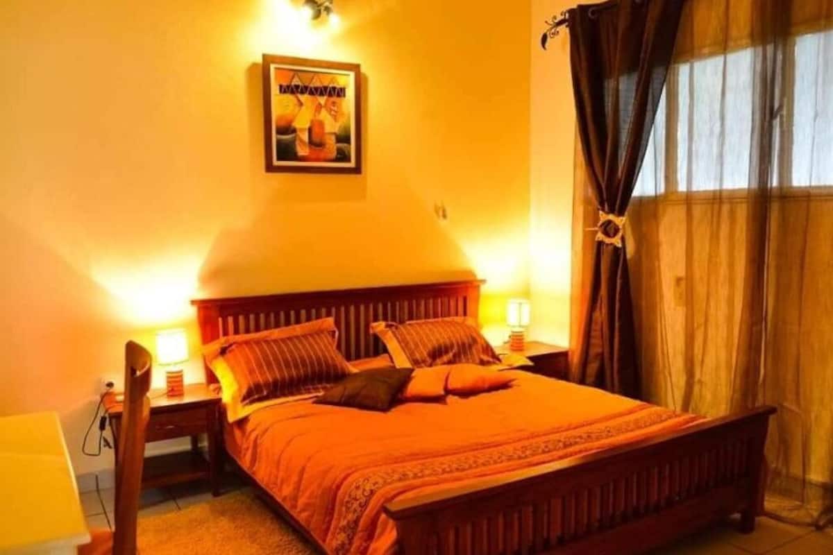 A bedroom with a wooden bed, two bedside tables with lamps, a wall-mounted picture, and a window with curtains.