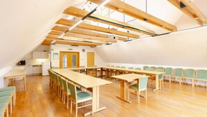Un salón de conferencias con mesas y sillas de madera, una kitchenette y un techo alto con vigas a la vista.