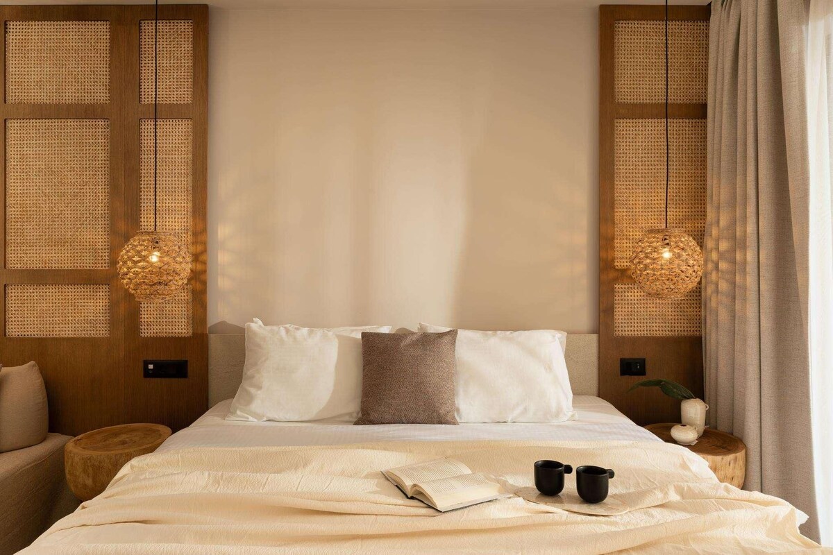 A neatly made bed with a book and two mugs, a wooden headboard, and two woven wall panels with hanging lamps.