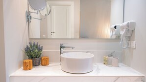 A bathroom with a round white sink, a mirror, a hairdryer, and a potted lavender plant.